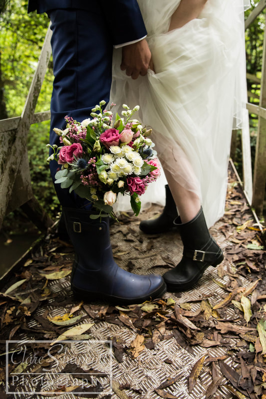Photograph of bride and groom on wedding day outside on abridge wearing boots as the groom holds the bride's dress up away from the floor.