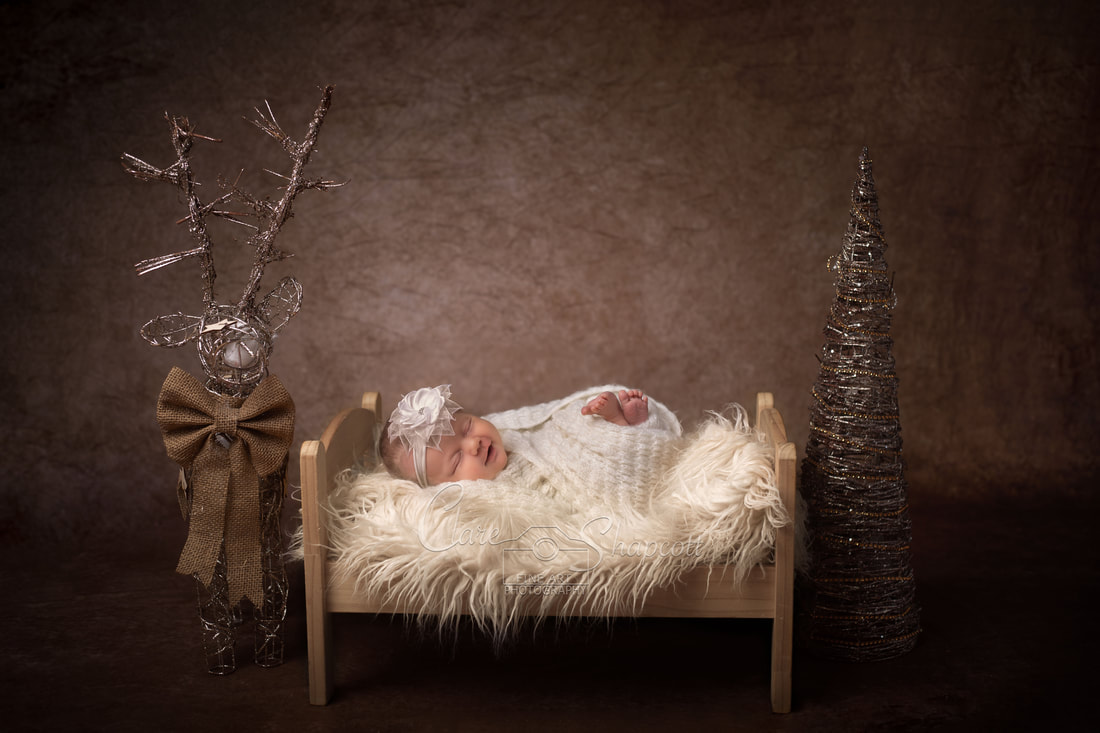 Newborn baby wrapped in soft white fabric sleeps on miniature bed with silver christmas tree and reindeer next to them.