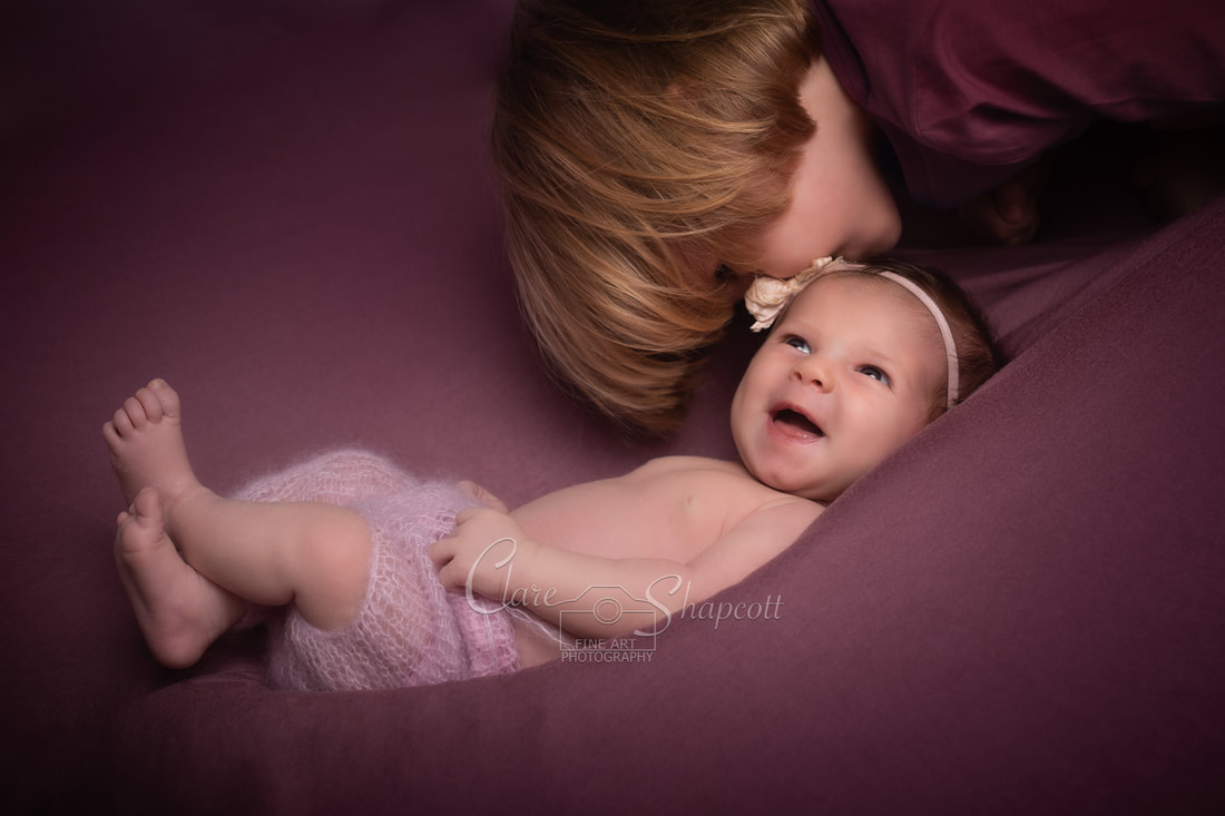 Happy newborn baby on pink backdrop with older sibling kissing on forehead