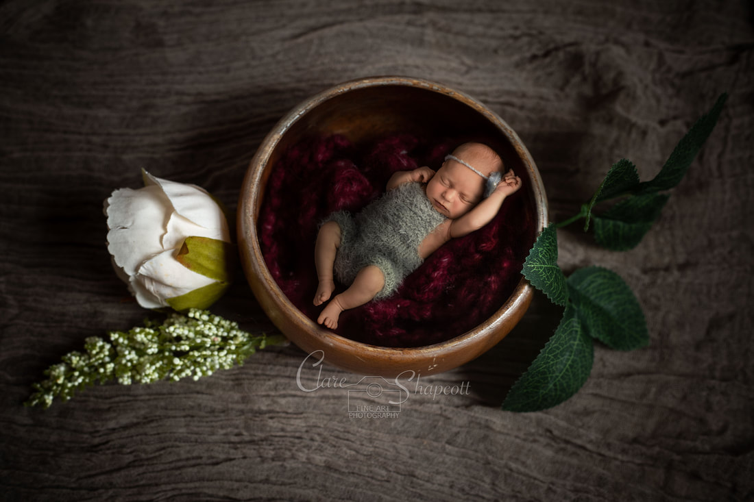 Sleeping newborn in grey knitted outfit lying in wooden bowl ontop of purple fabric next to green plants.
