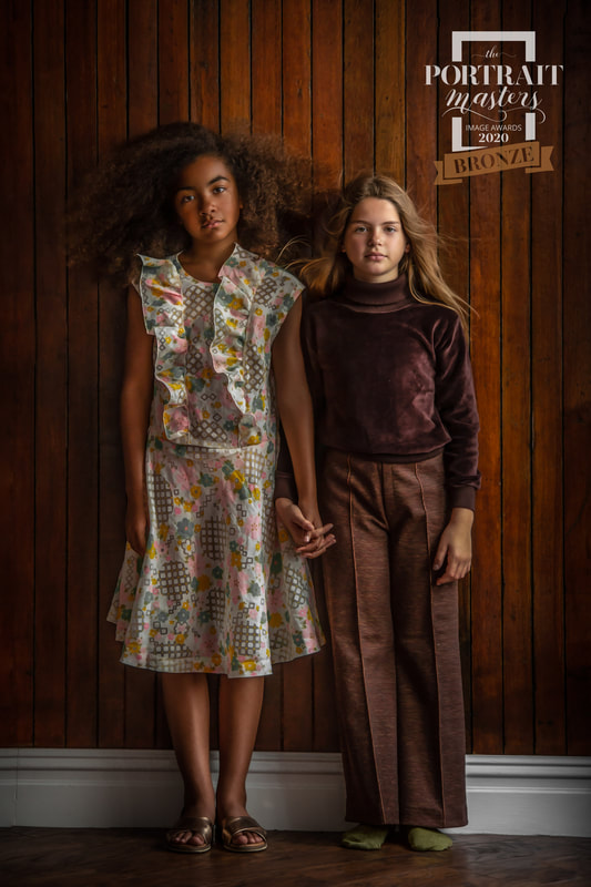 Two girls hold hands in front of brown wooden background in fashionable clothing.