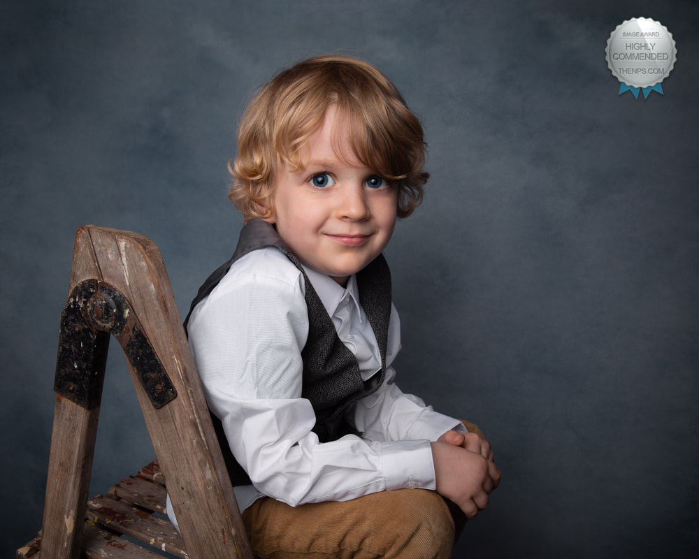 Fine art portrait of young smartly dressed boy sitting on step ladder with arms resting on knees.
