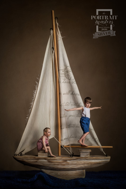 Fine art portrait of two young siblings sailing on toy wooden boat on top of deep blue material ocean.